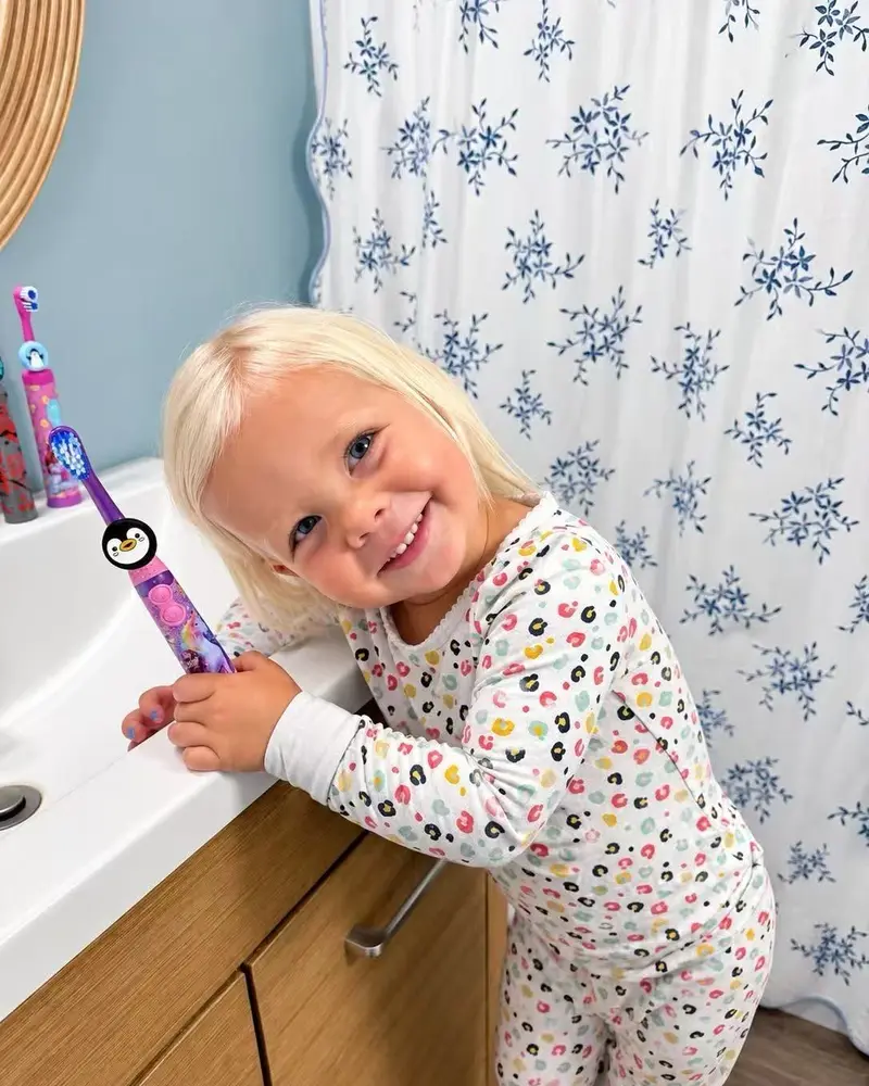 Smiling toddler at the sink holding a toothbrush with a penguin-skin Truthbrush Tracker